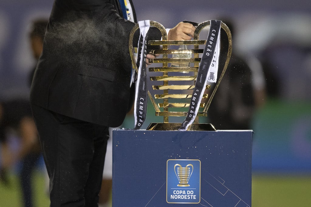 Taça da Copa do Nordeste sobre base com logo da competição e homem vestindo terno preto segurando alça do troféu