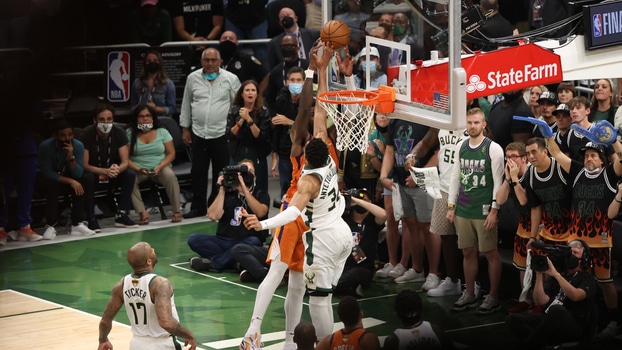 Jogadores de basquete em quadra, durante jogo. Dois deles pulando alto, junto à cesta, um tentando evitar que o outro pontue.