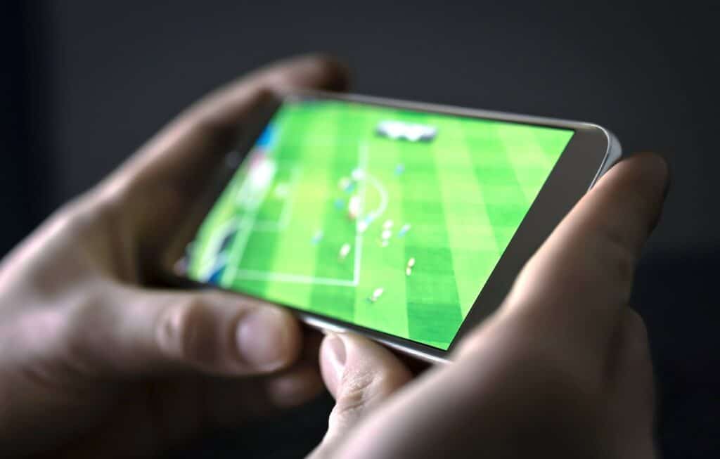 Mãos segurando um celular na horizontal e, na tela, um jogo de futebol está passando
