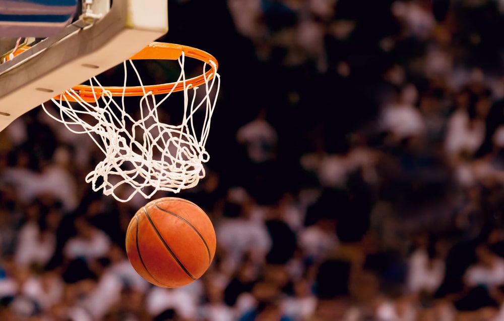 Bola de basquete passando por cesta e, de fundo em desfoque, torcedores acompanhando o jogo