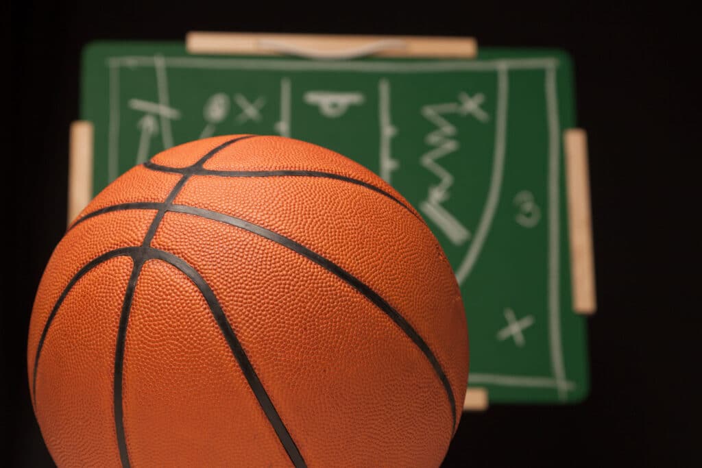 Bola de basquete em foco. No fundo, quadro tático de treinador de basquete, com rabiscos de estratégia de jogo, em desfoque.