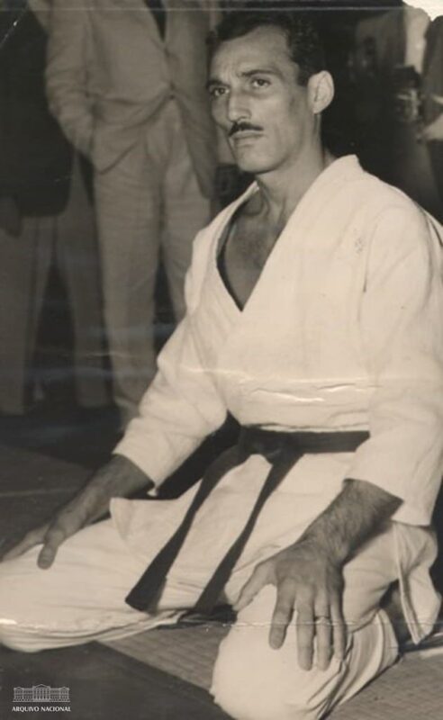 Foto em preto e branco, de Hélio Gracie jovem, vestindo kimono de jiu-jitsu, ajoelhado no tatame
