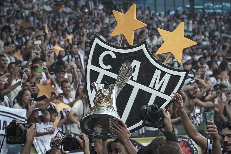 Taça de campeão da Série A do Campeonato Brasileiro de 2021 erguida por integrante do elenco do Atlético-MG, várias câmeras fotográficas e o escudo do Galo atrás da taça. No fundo, a torcida do Atlético-MG.
