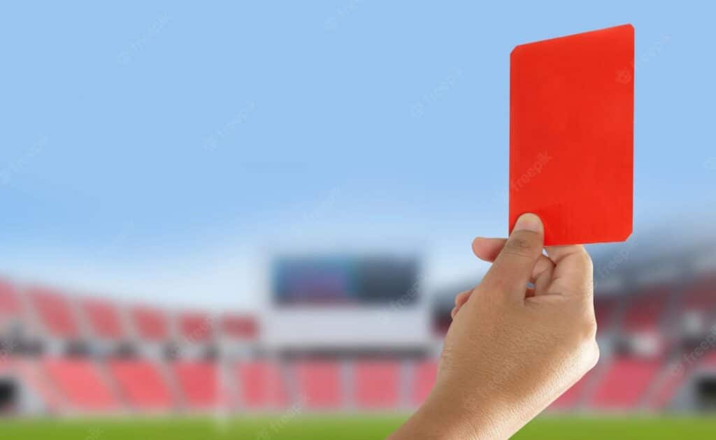 Mão segurando cartão vermelho. De fundo, estádio de futebol em desfoque