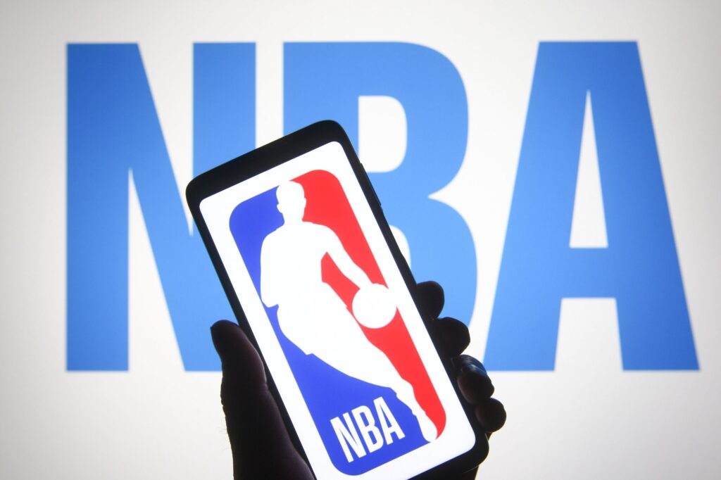 Mão de uma pessoa segurando um celular e, na tela do aparelho, a logo da NBA. No fundo, em letras azuis sobre fundo branco, as letras NBA.