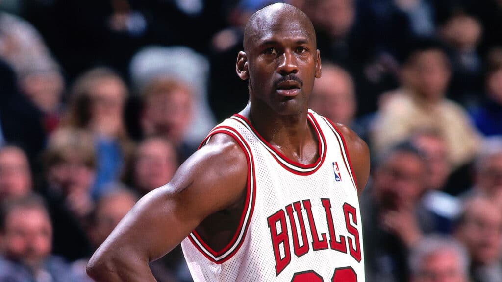 Michael Jordan, vestindo camisa branca e vermelha dos Bulls, em quadra. De fundo, torcida aparece desfocada.