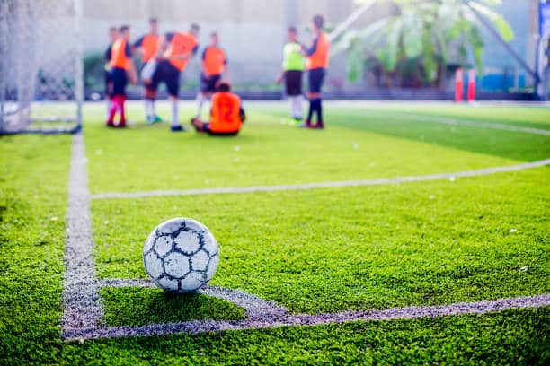 Bola de futebol na marca do escanteio, linhas do gramado marcadas e jogadores ao fundo, na pequena área, esperando cobrança do escanteio