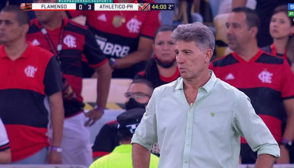 Renato Gaúcho quando era técnico do Flamengo, aparece com expressão de insatisfação, em imagem de TV. No canto esquerdo superior, o placar: Flamengo 0x3 Athletico-PR e, ao fundo, torcedores do Flamengo.