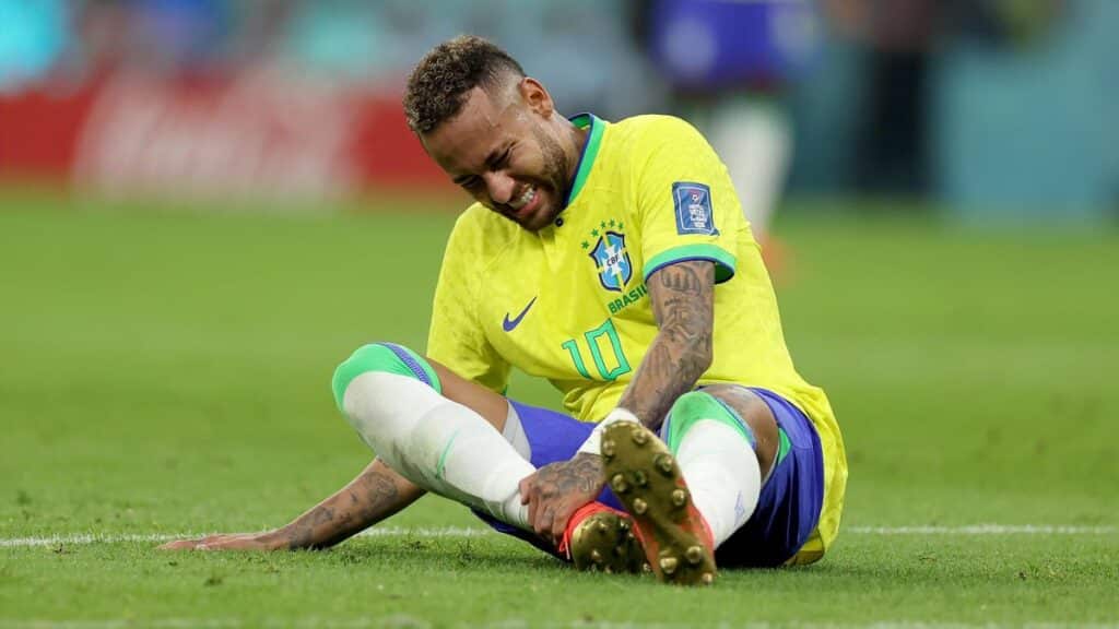Neymar, jogador de futebol, sentado no gramado com as mãos no tornozelo, após se lesionar na Copa do Mundo de 2022
