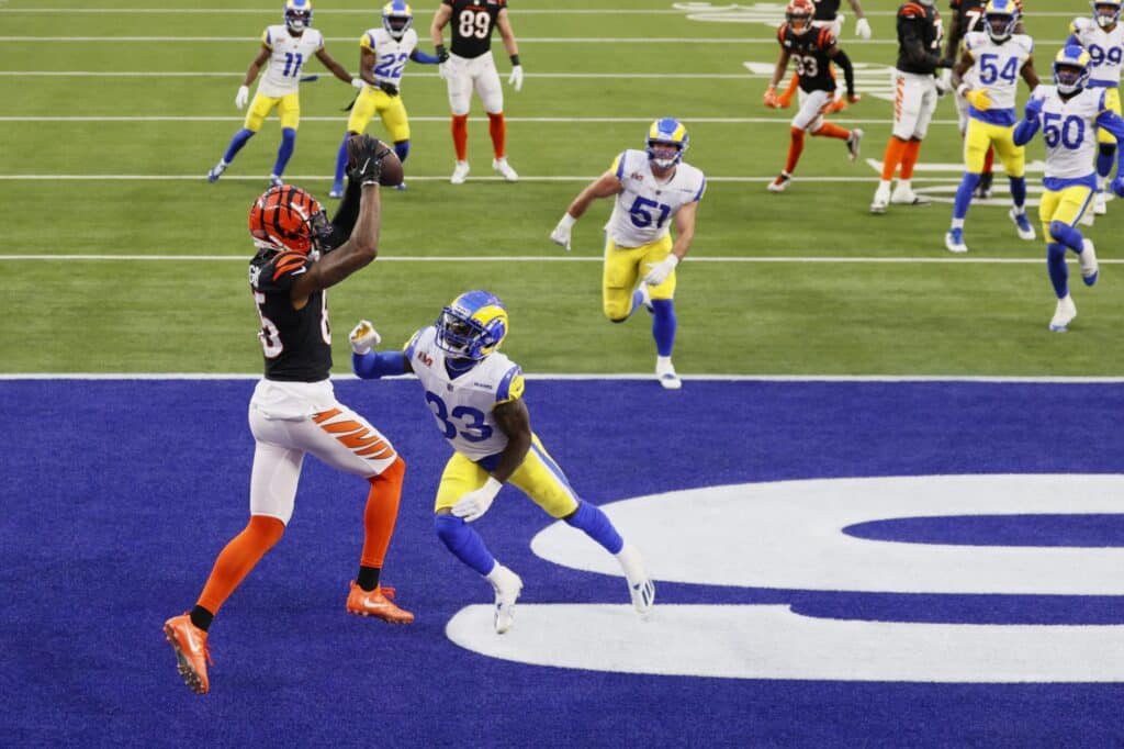 Jogador recebendo a bola na endzone, enquanto um jogador adversário tenta interceptá-lo; ao fundo outro jogador entrando na endzone e mais jogadores observando a jogada